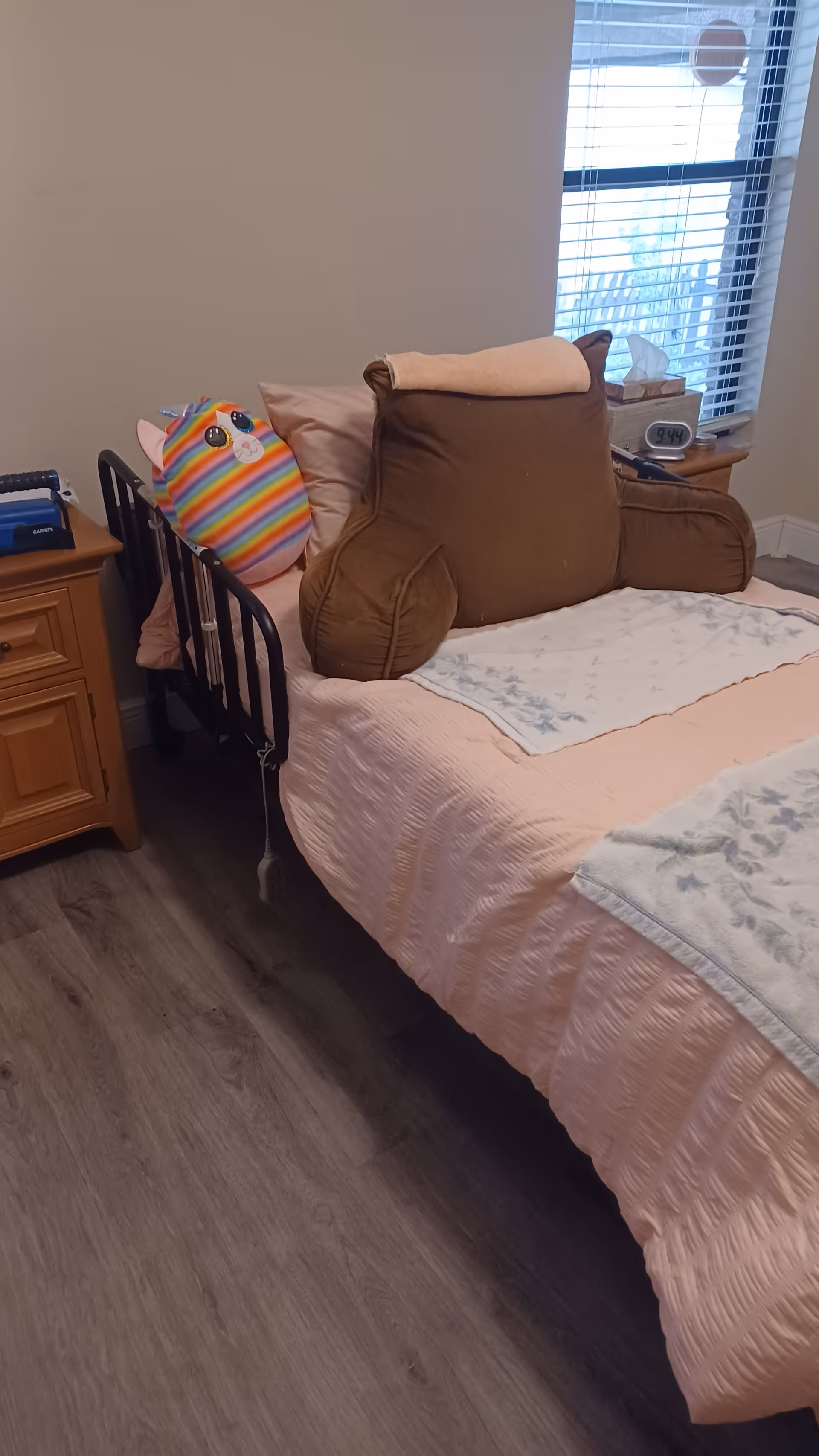 A bed with pink quilt, a large brown stuffed pillow, and a nightstand beside a window.