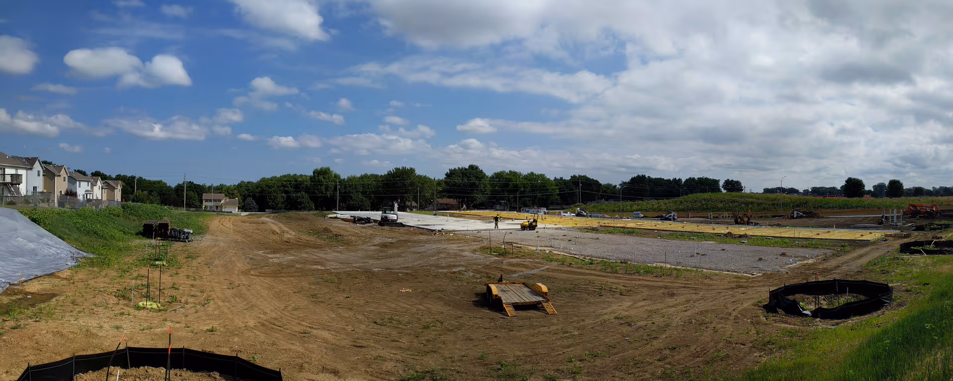 Wide panoramic view of a construction site with cleared land and foundation work in progress under a partly cloudy sky. There are some houses on the left side and trees in the background.