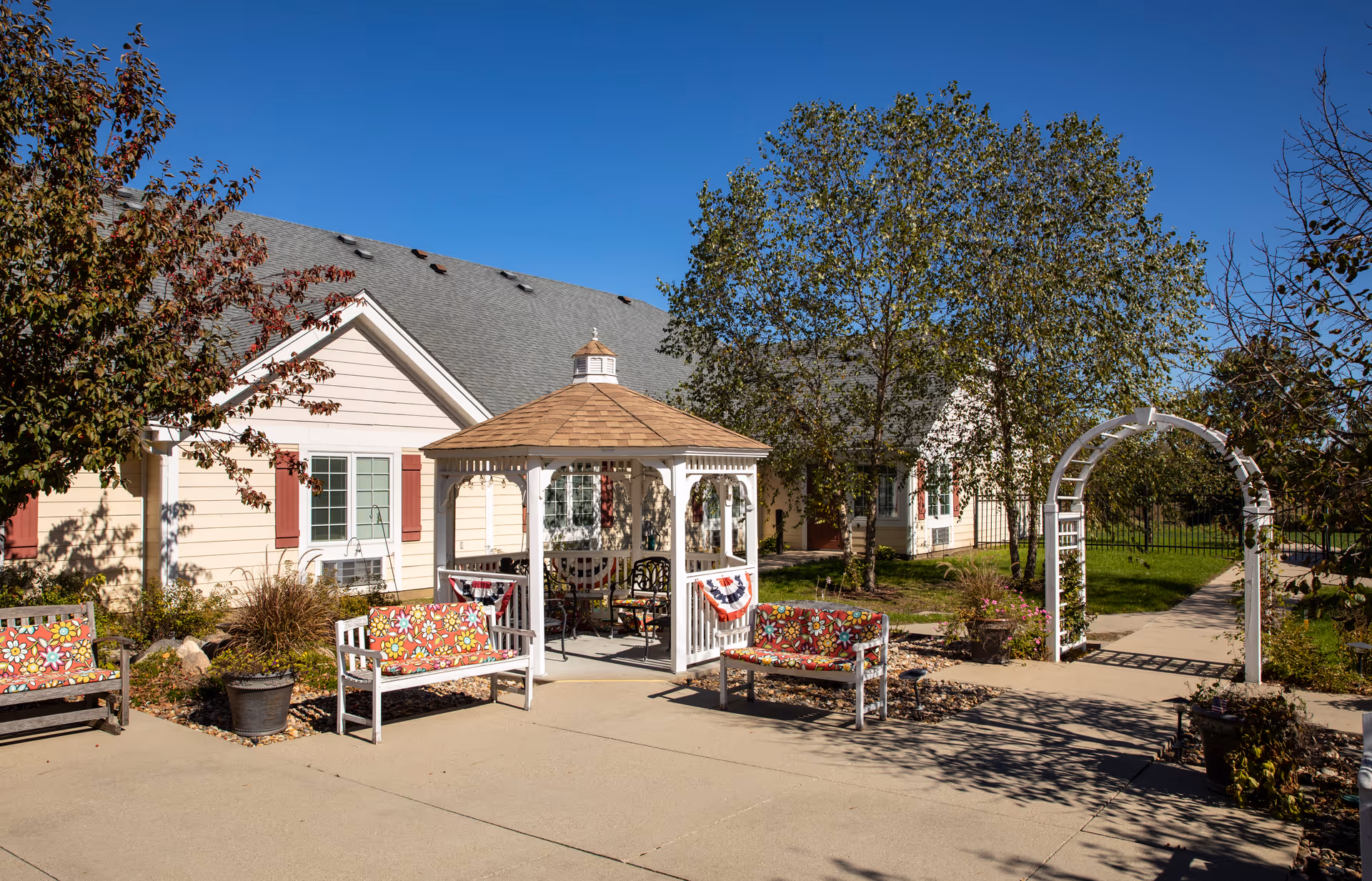 Outdoor courtyard in front of a beige assisted living building with a white gazebo, colorful benches, and a walkway under a clear blue sky.