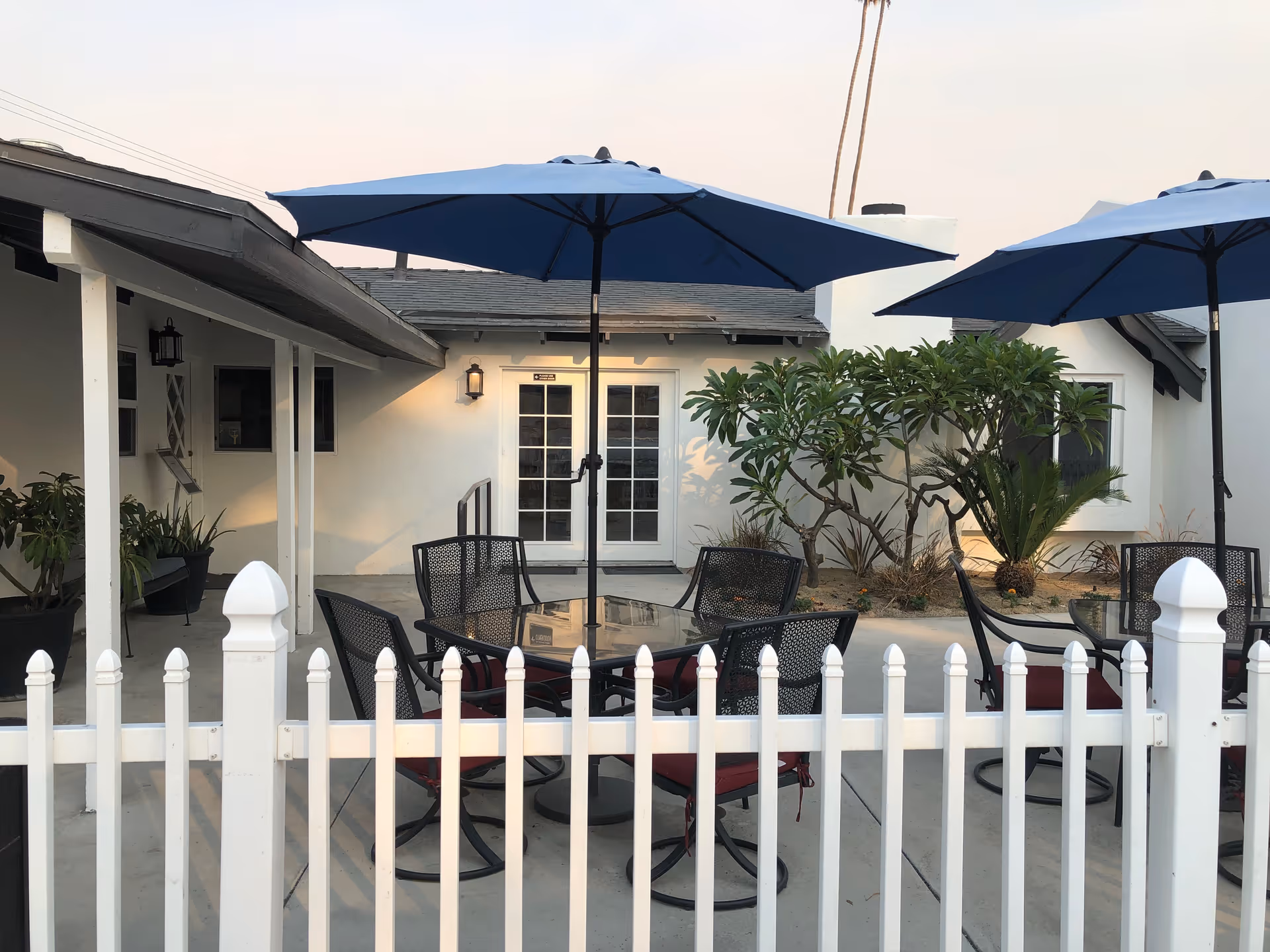 Outdoor patio with glass-top tables, metal chairs, and blue umbrellas in front of a white building.