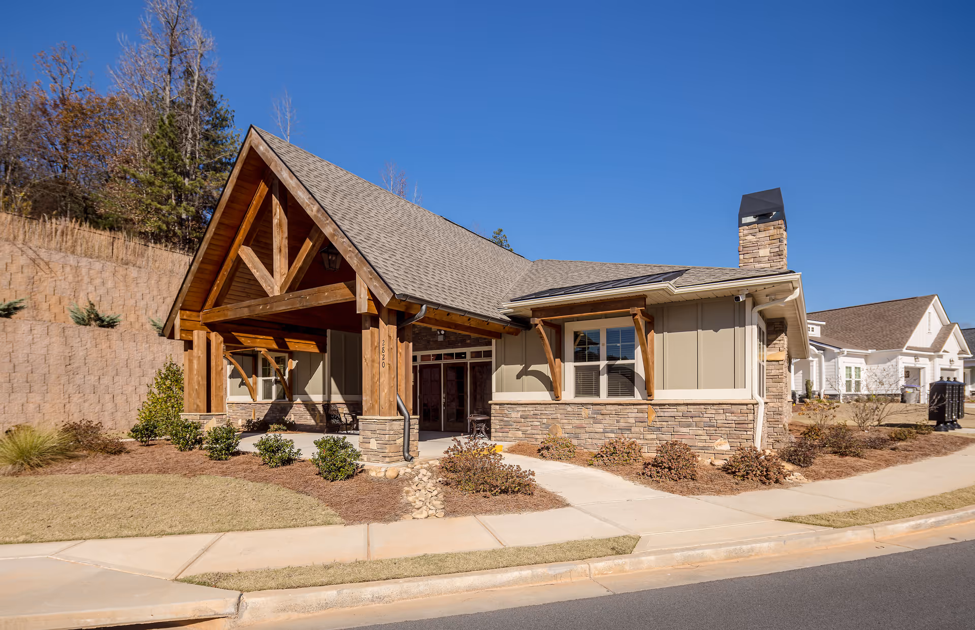 Exterior view of a single-story building with a peaked roof and wooden beams at the entrance, surrounded by landscaped bushes and a sidewalk under a clear blue sky.