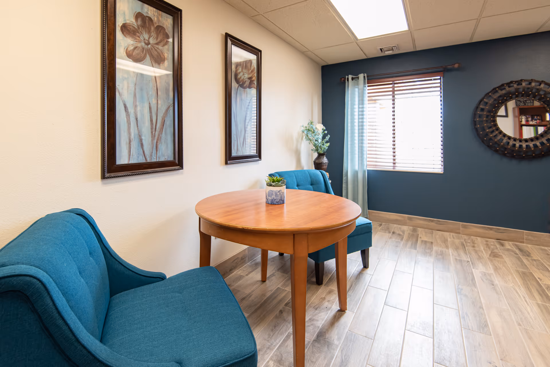 Small sitting area with teal upholstered chairs and a round wooden table beneath framed floral artwork and a window.