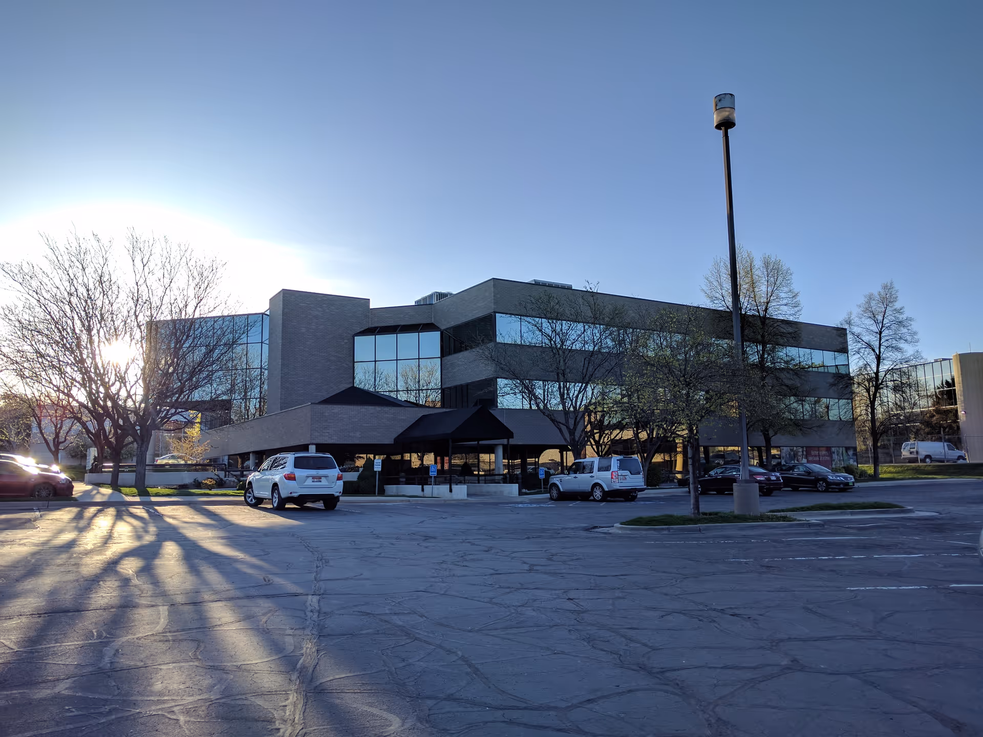 Modern glass-front building with a covered entrance and parked cars in the lot.