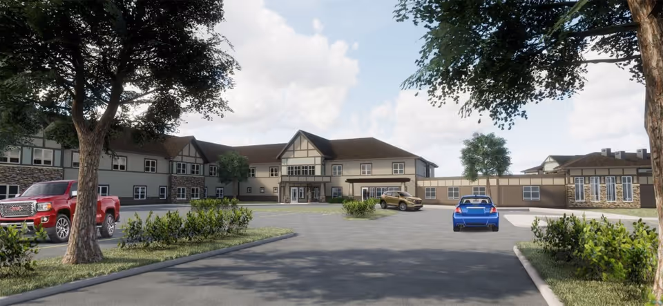 Front exterior view of Prairie Pointe Assisted Living facility with a parking lot in front, several parked cars, trees, and shrubs under a partly cloudy sky.