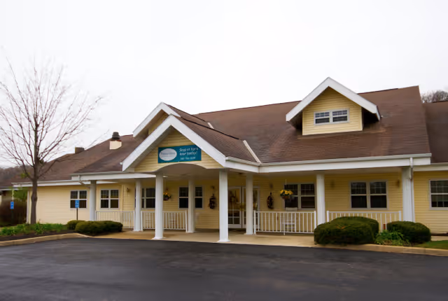 Single-story yellow senior living building with a covered front entrance, white columns, and a small parking area.