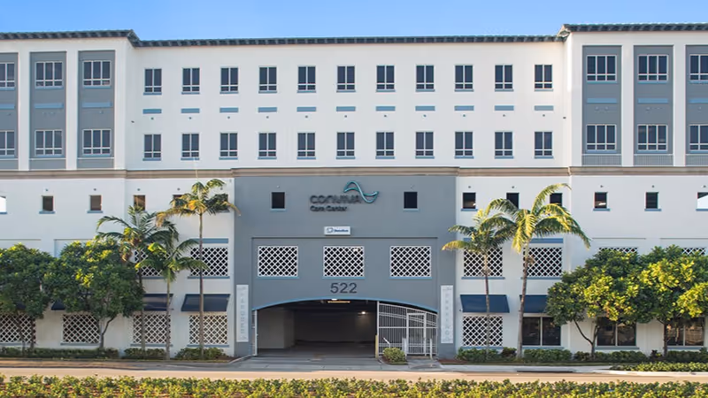 Front view of a multi-story senior living facility with palm trees and an archway.