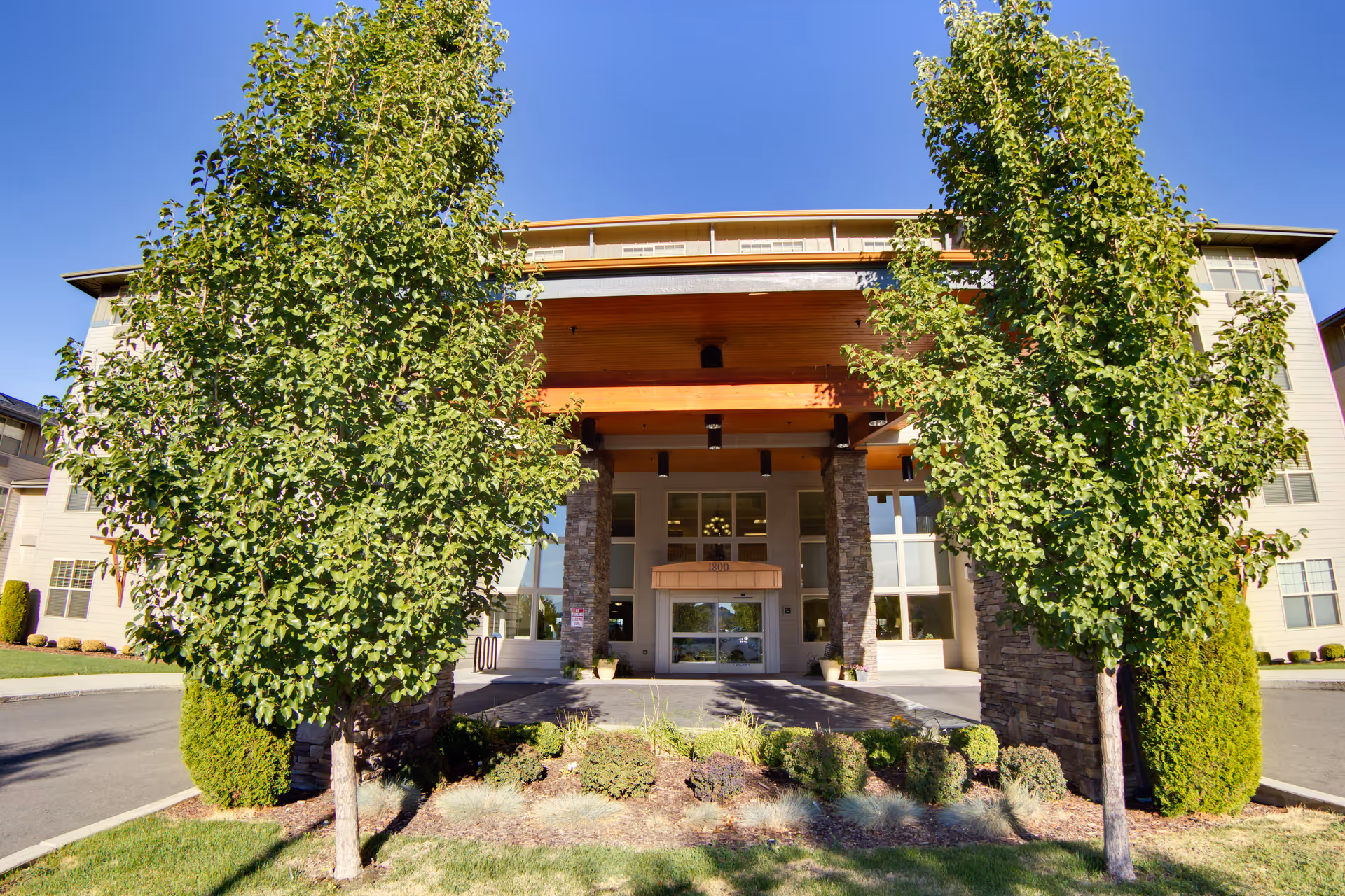 Front exterior view of Bonaventure of Tri-Cities building with a covered entrance supported by stone pillars, flanked by two tall green trees and landscaping in front under a clear blue sky.