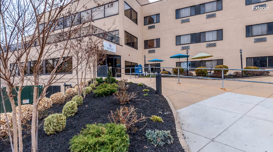 Exterior view of Serenity Rehabilitation And Health Center showing the building entrance, landscaped garden with bushes and small trees, a paved walkway with a ramp, and outdoor seating with umbrellas.