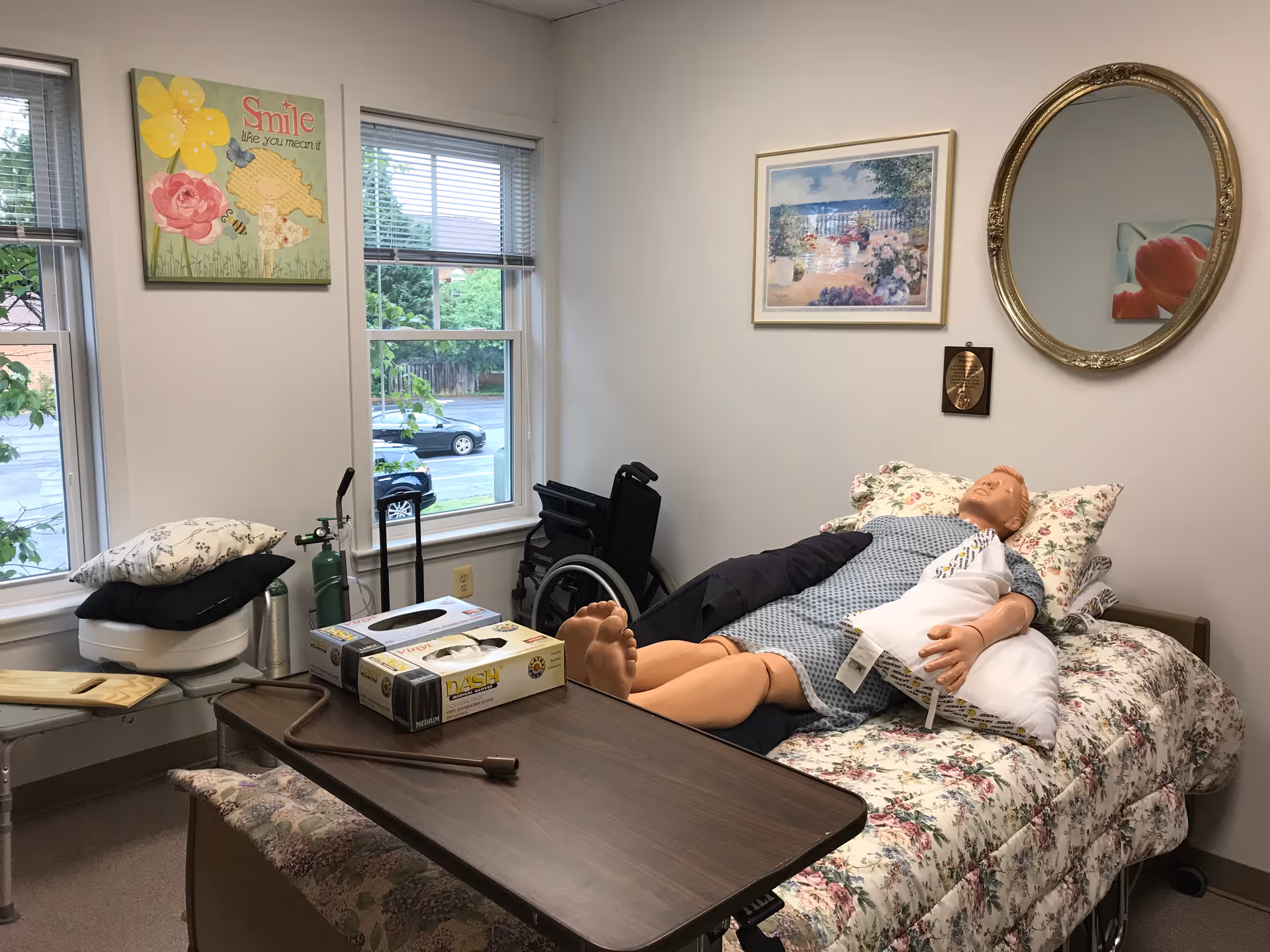 Interior of a senior care room with a bed, a medical mannequin, wheelchair, and supplies.