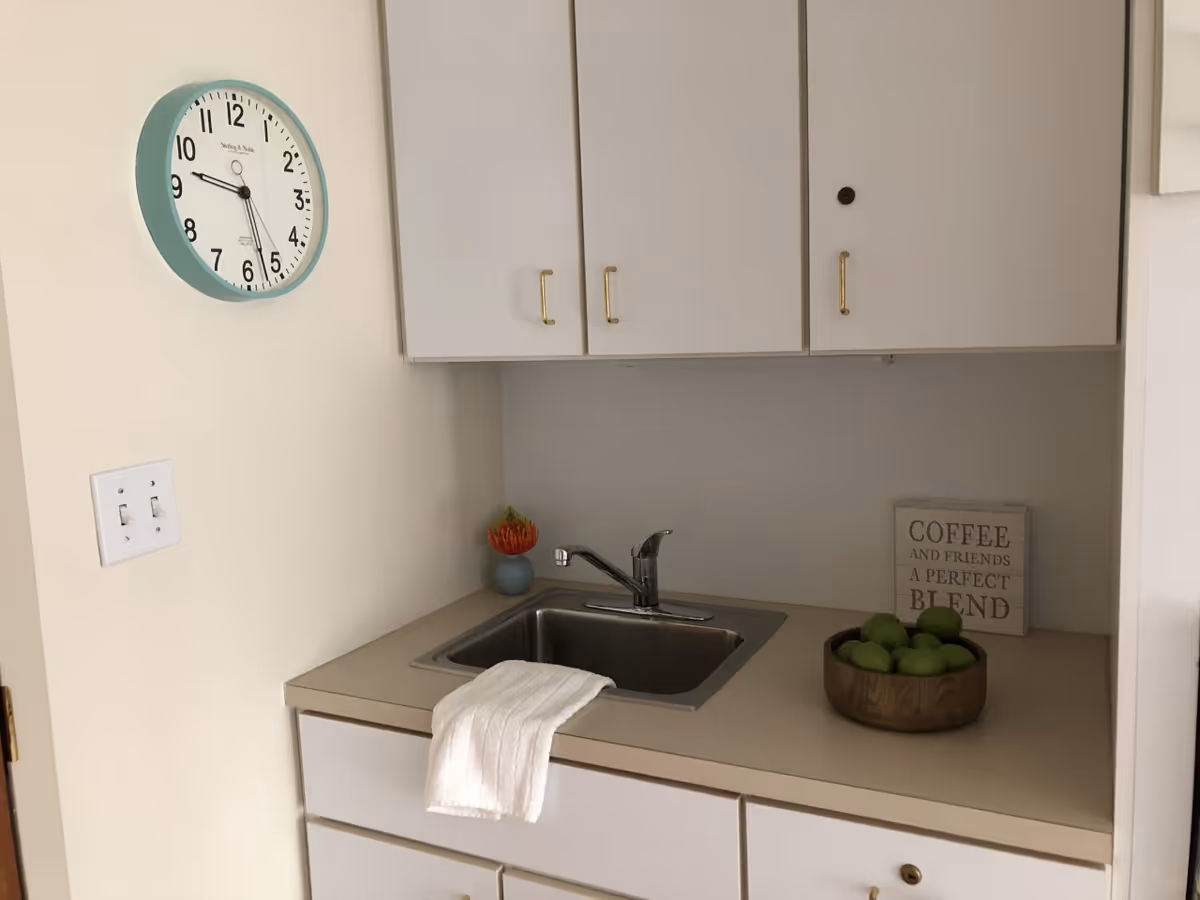 A small kitchen area with a stainless steel sink, white cabinets with gold handles, a white towel hanging over the sink edge, a bowl of green apples on the countertop, a small potted plant, and a decorative sign that reads 'COFFEE AND FRIENDS A PERFECT BLEND'. A round wall clock with a teal frame shows the time as 9:28.