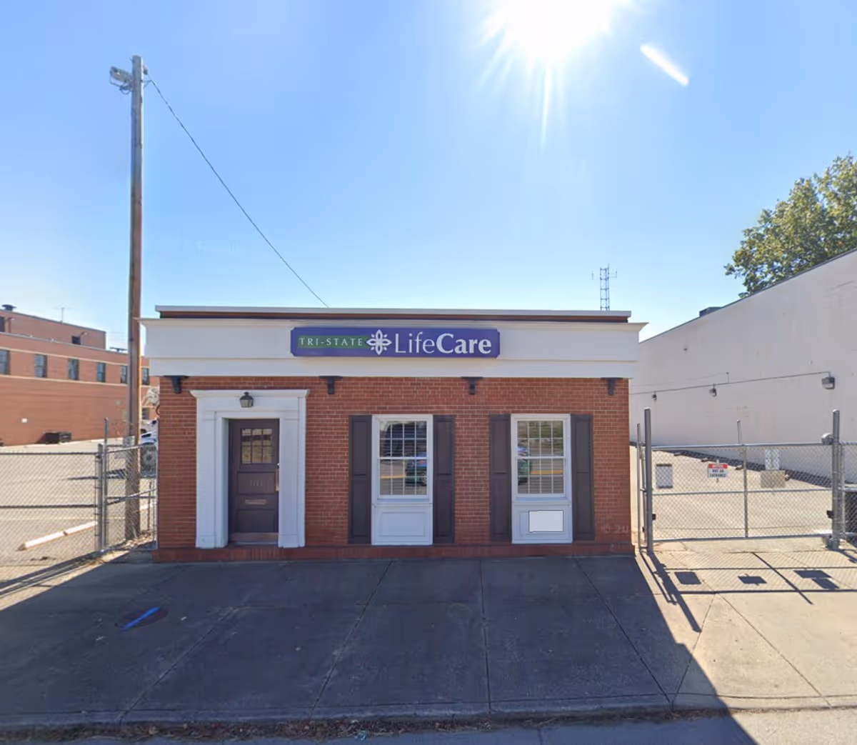 Small brick building with Tri-State LifeCare sign, fenced lot, and bright sunny sky.