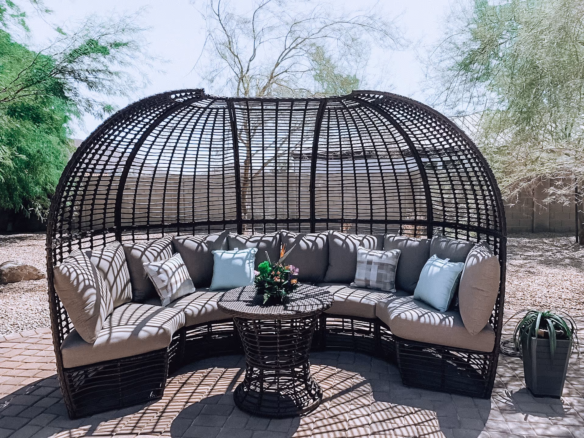 A dome-shaped wicker outdoor sofa with cushions surrounding a round wicker table on a paved patio under trees.