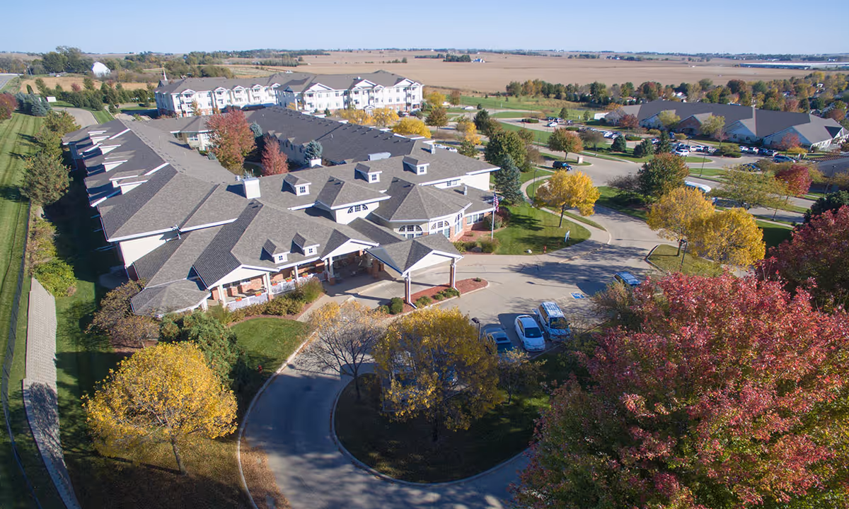 Aerial view of a multi-wing senior living building with a covered entrance, circular driveway, parked cars and landscaped grounds in autumn.