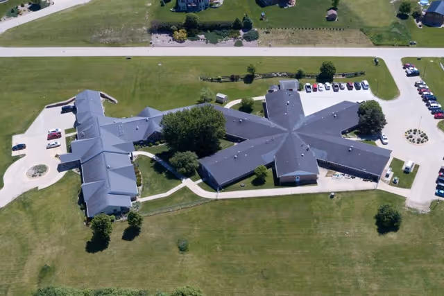 Aerial view of Mica Hill Estates showing a large building complex with multiple wings extending from a central area, surrounded by green lawns, trees, parking lots with cars, and paved walkways.