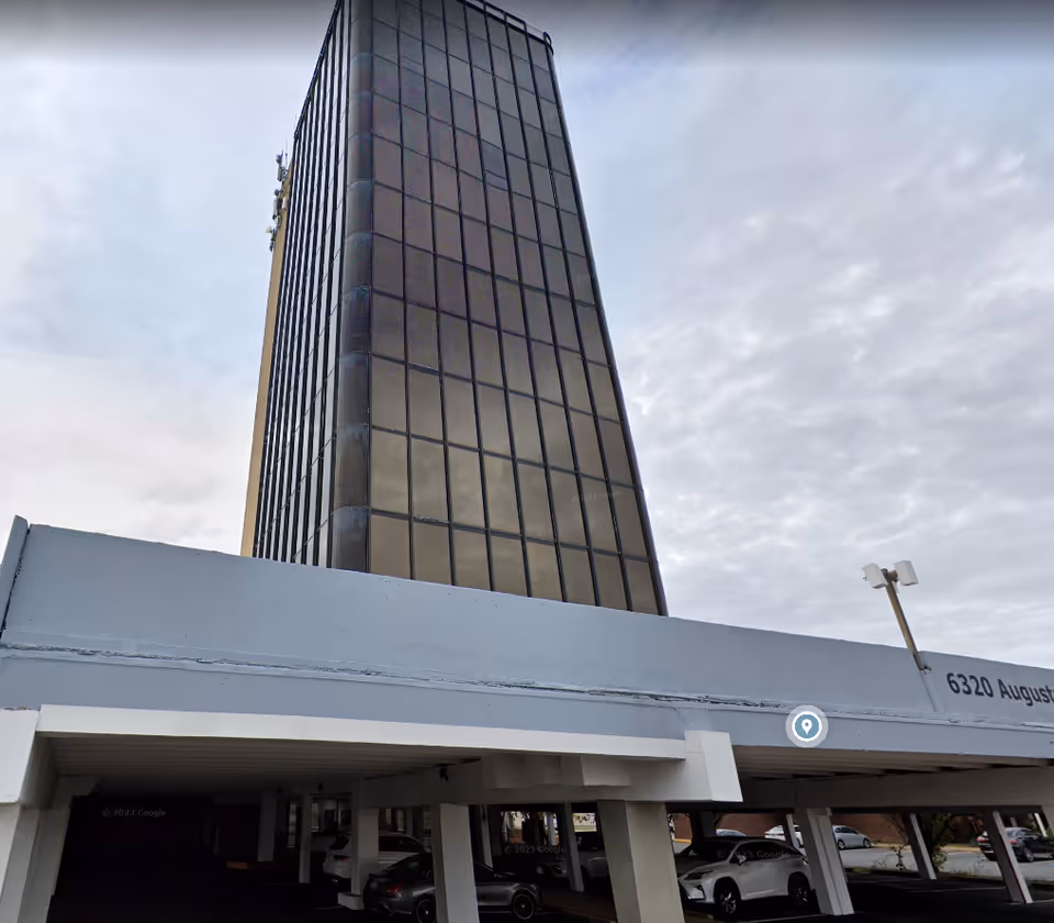 Tall, modern building exterior with a parking area beneath and a cloudy sky.