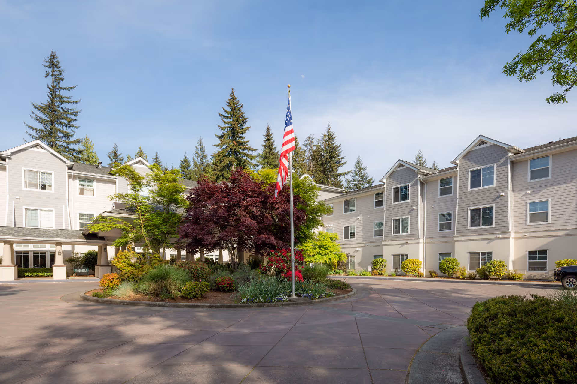 Exterior view of Cogir of Mill Creek senior living facility showing a circular driveway with landscaped greenery and an American flag in the center, surrounded by a three-story building with multiple windows under a clear blue sky.