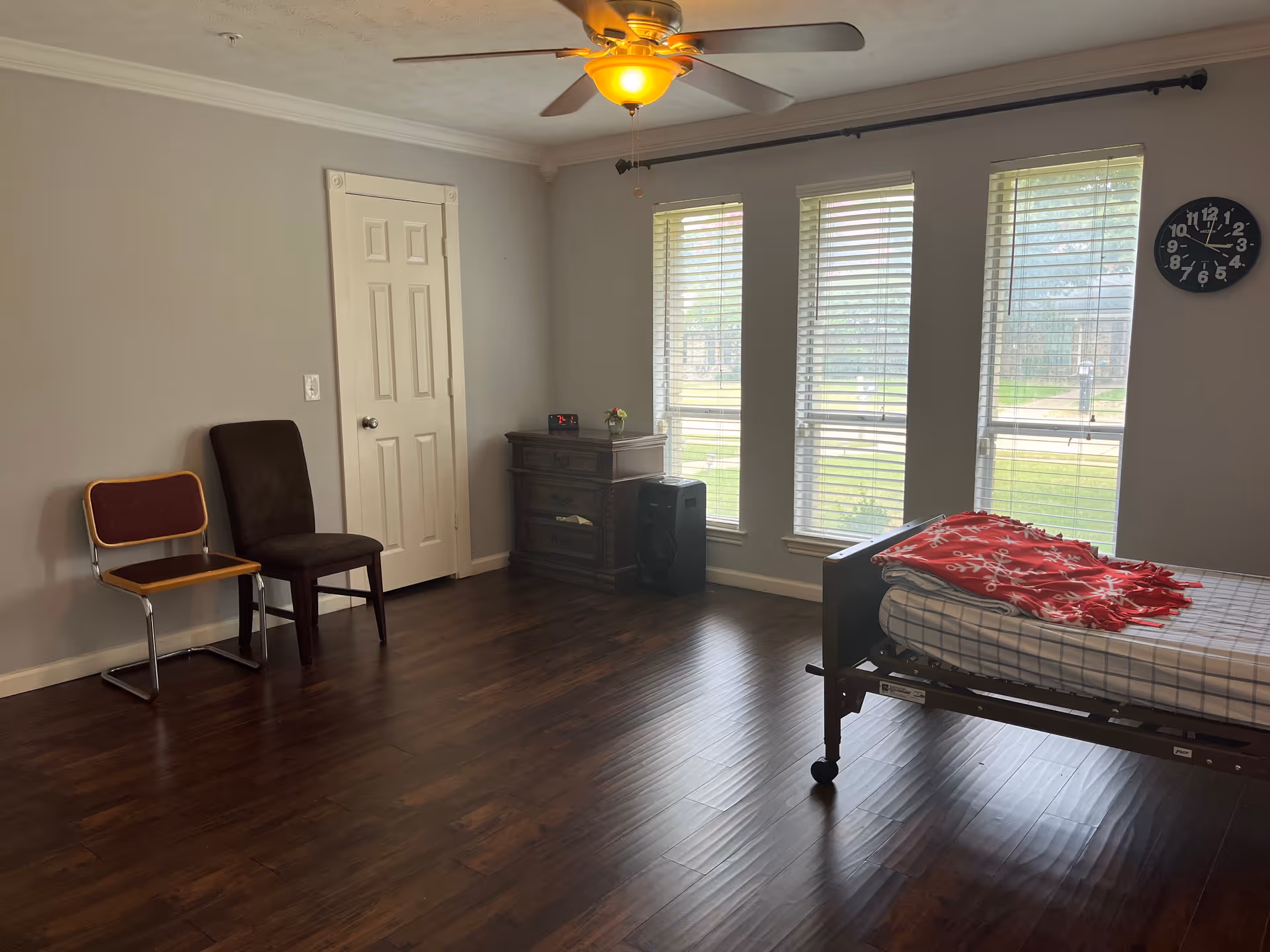 A simple bedroom in a care home with a bed, dresser, chairs, and windows.