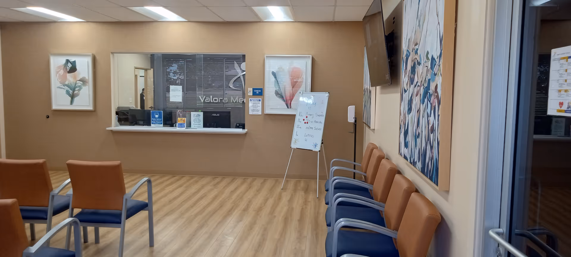 Interior waiting area with orange and blue chairs arranged along the wall near a reception window.