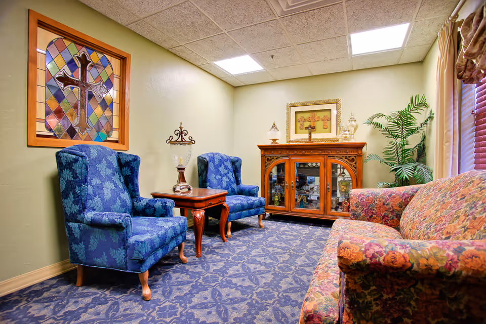 Cozy living room at Grace Living Center-Bethany with blue chairs, floral sofa, and cabinet.