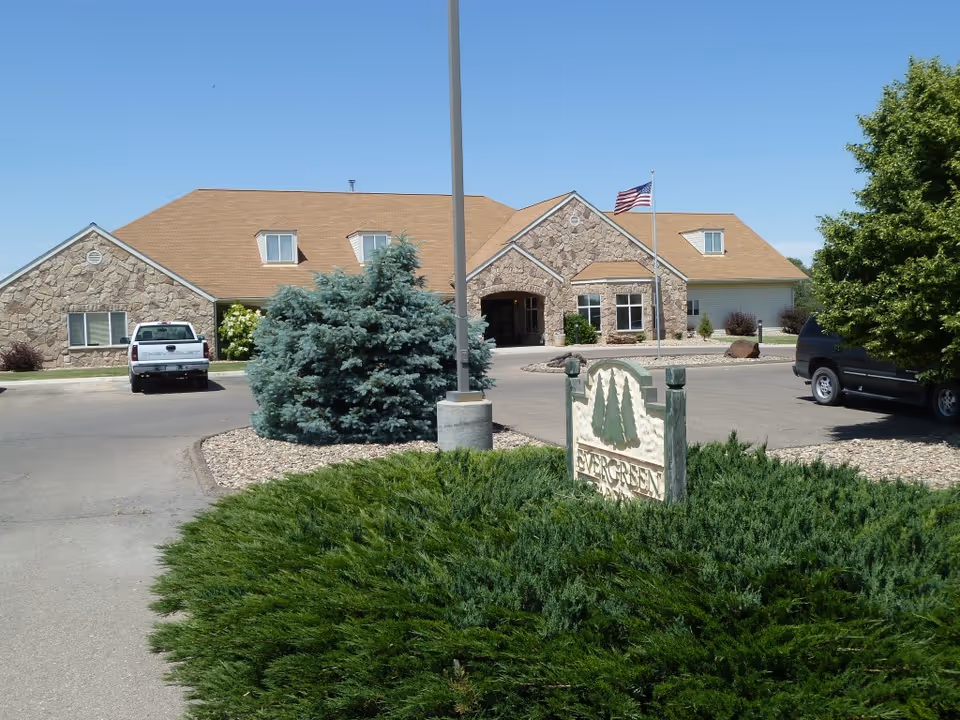 Exterior view of a single-story stone and siding building with a tan roof, an American flag on a flagpole, two parked vehicles, and green shrubs and trees in the foreground. A sign reading 'Evergreen' is partially visible among the greenery.
