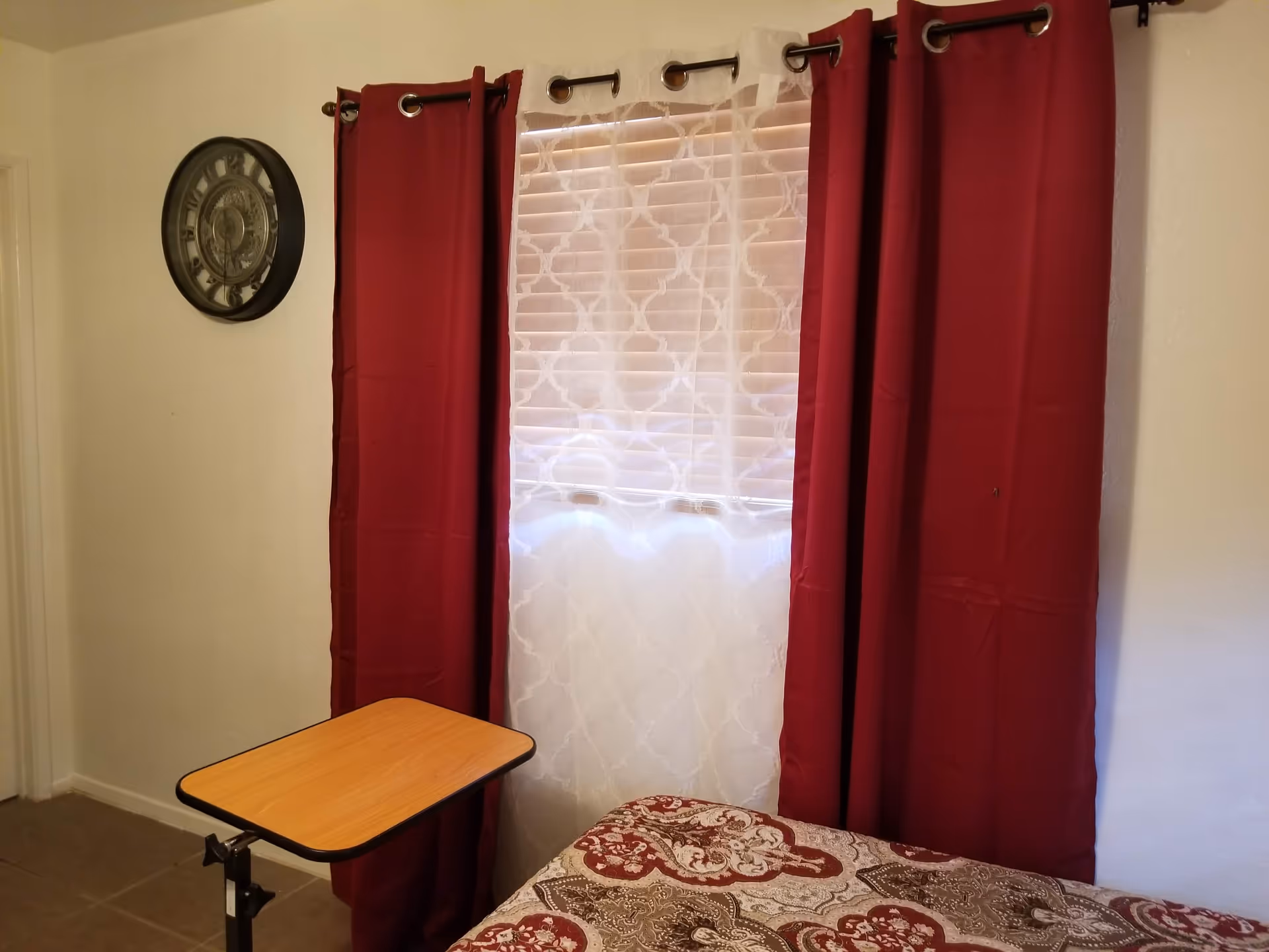 A small bedroom with red curtains, a bed, a wooden overbed table, and a wall clock.