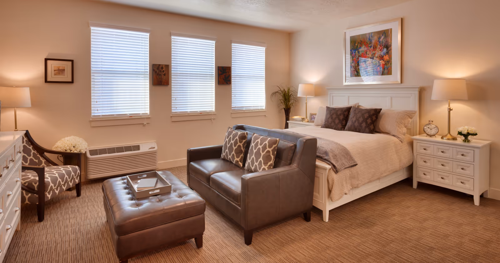 Well-lit furnished bedroom with a bed, loveseat, ottoman, armchair, nightstands and three windows with blinds.