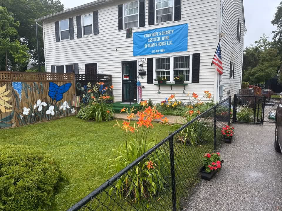 White two-story building with a blue banner, garden, flowers, and a black chain-link fence.