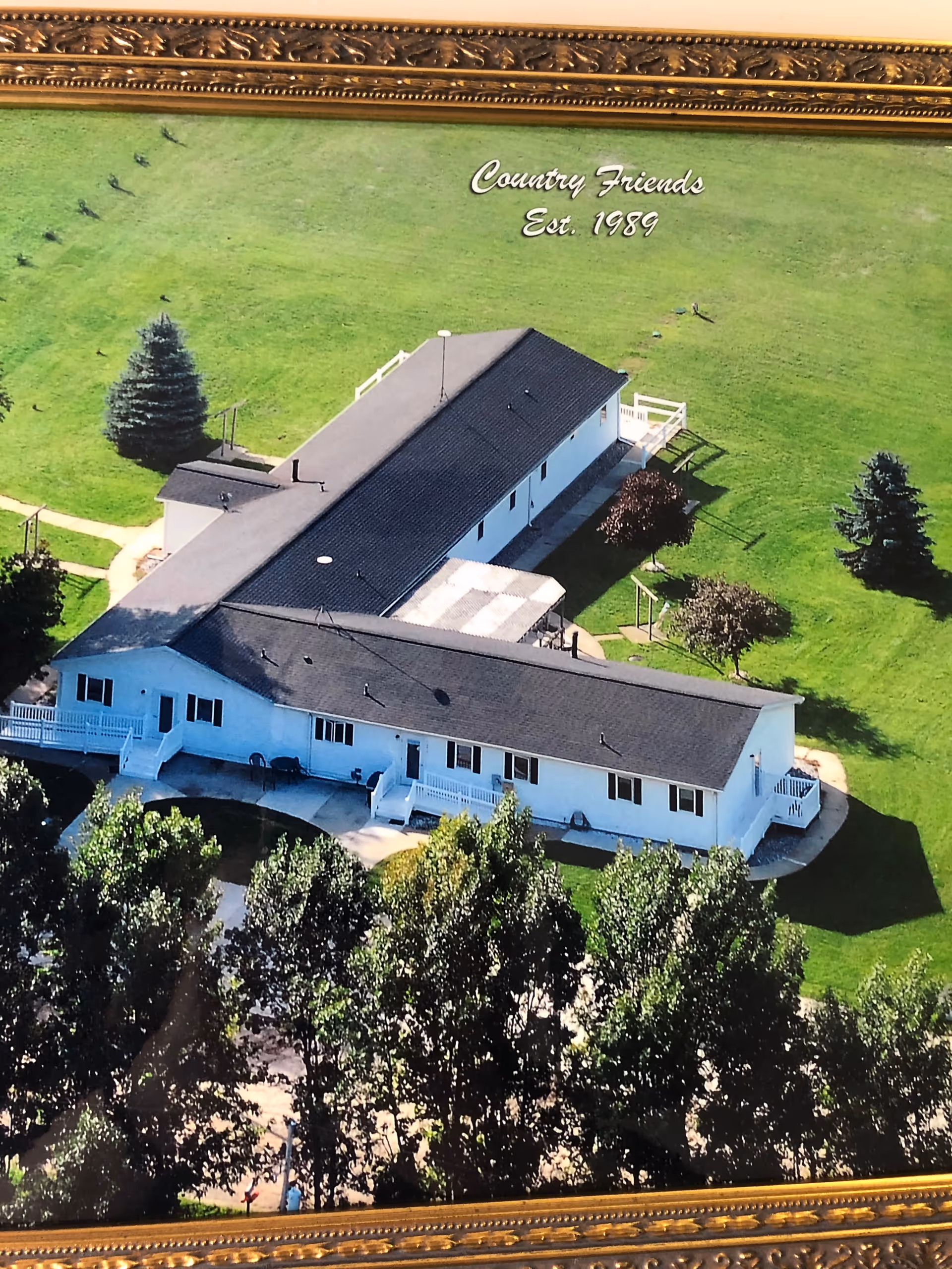 Aerial view of a single-story white building with a dark roof surrounded by green grass and trees. The building has multiple entrances with small staircases and a paved walkway. Text on the image reads 'Country Friends Est. 1989'.