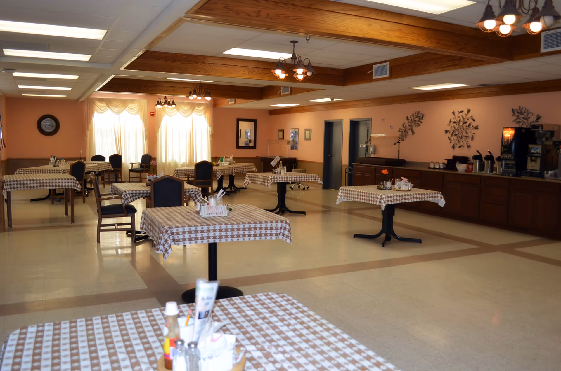 Spacious dining room with several tables covered in checkered tablecloths, chairs, wood-beamed ceiling, and a buffet counter along the right wall.