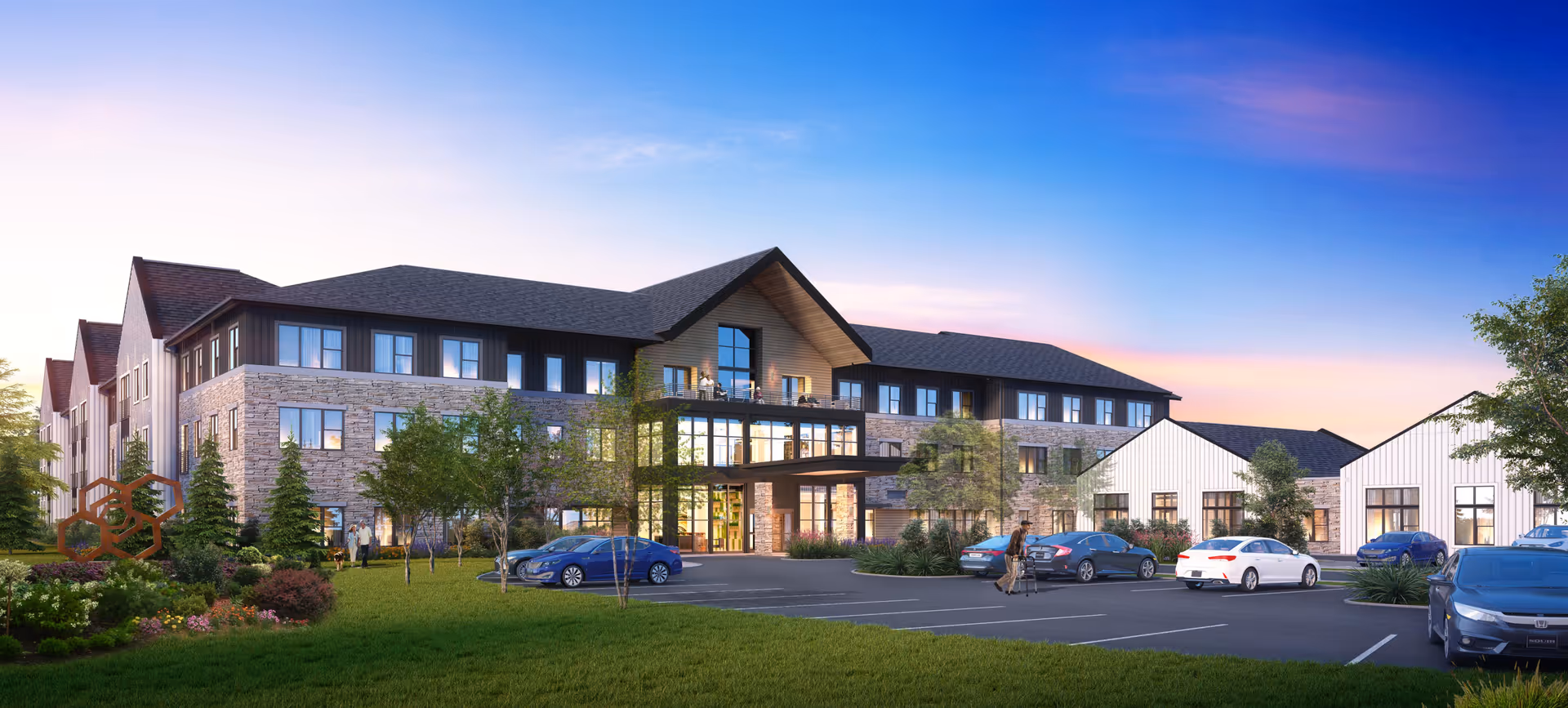 Front exterior of a three-story senior living building with landscaped grounds, parked cars, and a lit entrance at sunset.
