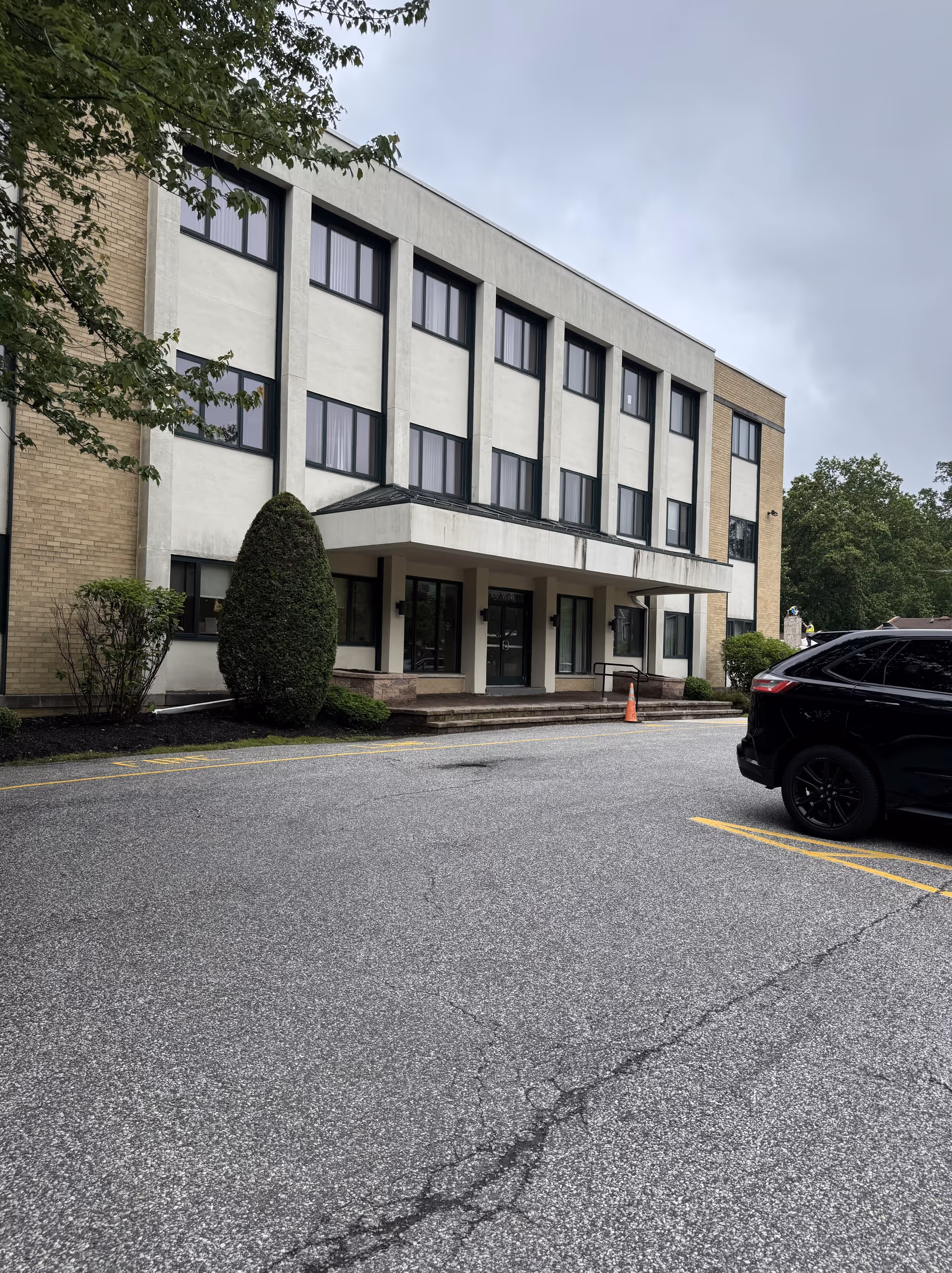 Three-story beige brick building with large windows, entrance canopy, and parked SUV in foreground.