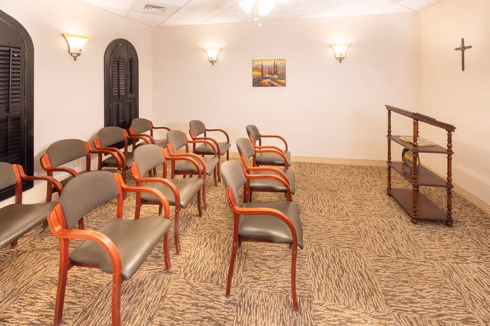 A small room with several rows of wooden chairs with padded seats and backs arranged facing a wooden lectern. The room has beige walls, carpeted floor with a patterned design, three wall-mounted light fixtures, a small painting on the wall, and a crucifix hanging on the right wall.