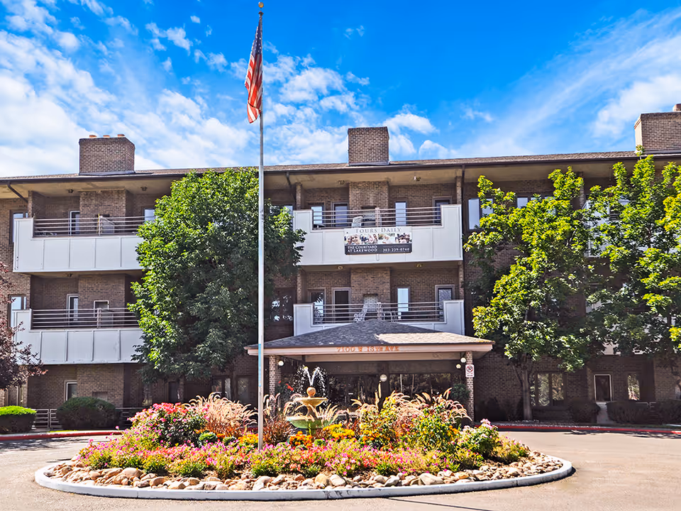 Front exterior of a multi-story brick senior living building with balconies, an American flagpole, and a landscaped fountain and flowerbed in front.