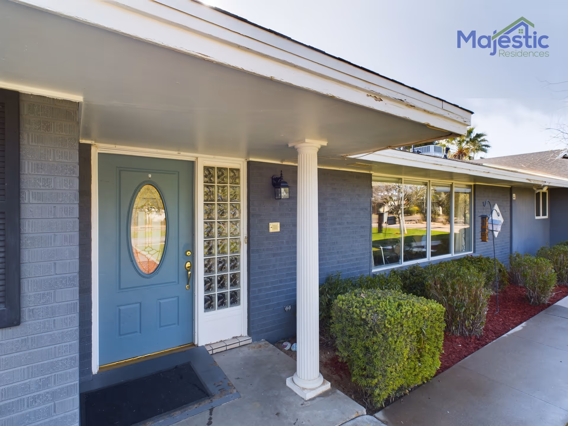 Blue front door with oval window, glass block panel, white pillar, and shrubs outside.