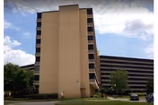 Exterior view of a tall tan senior living building with a central tower and parking.