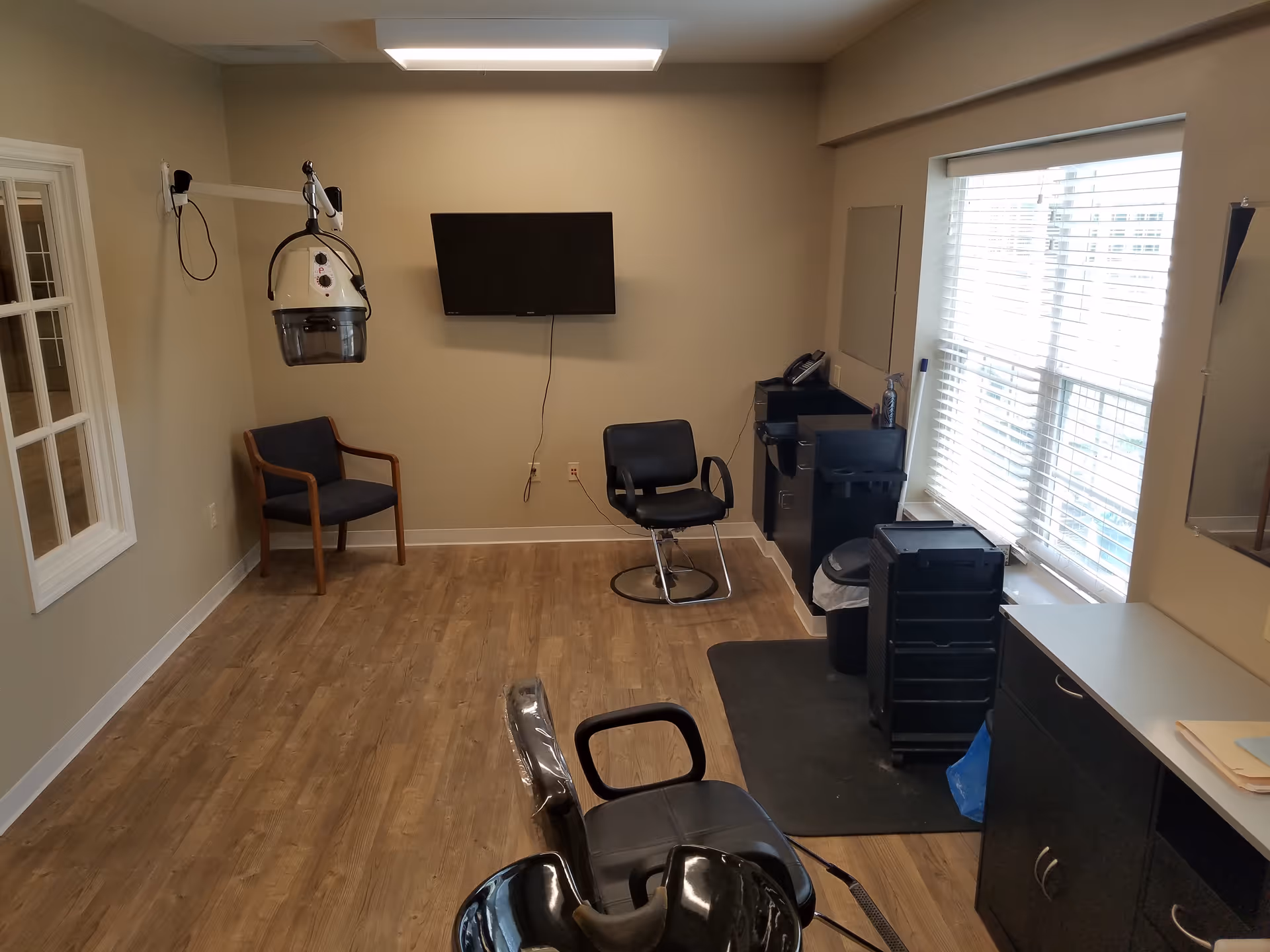 Interior salon room with a hooded dryer, chairs, a TV, and large window blinds.