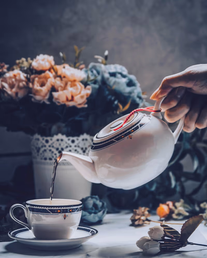 Hand pours hot tea from a teapot into a cup on a floral table setting.