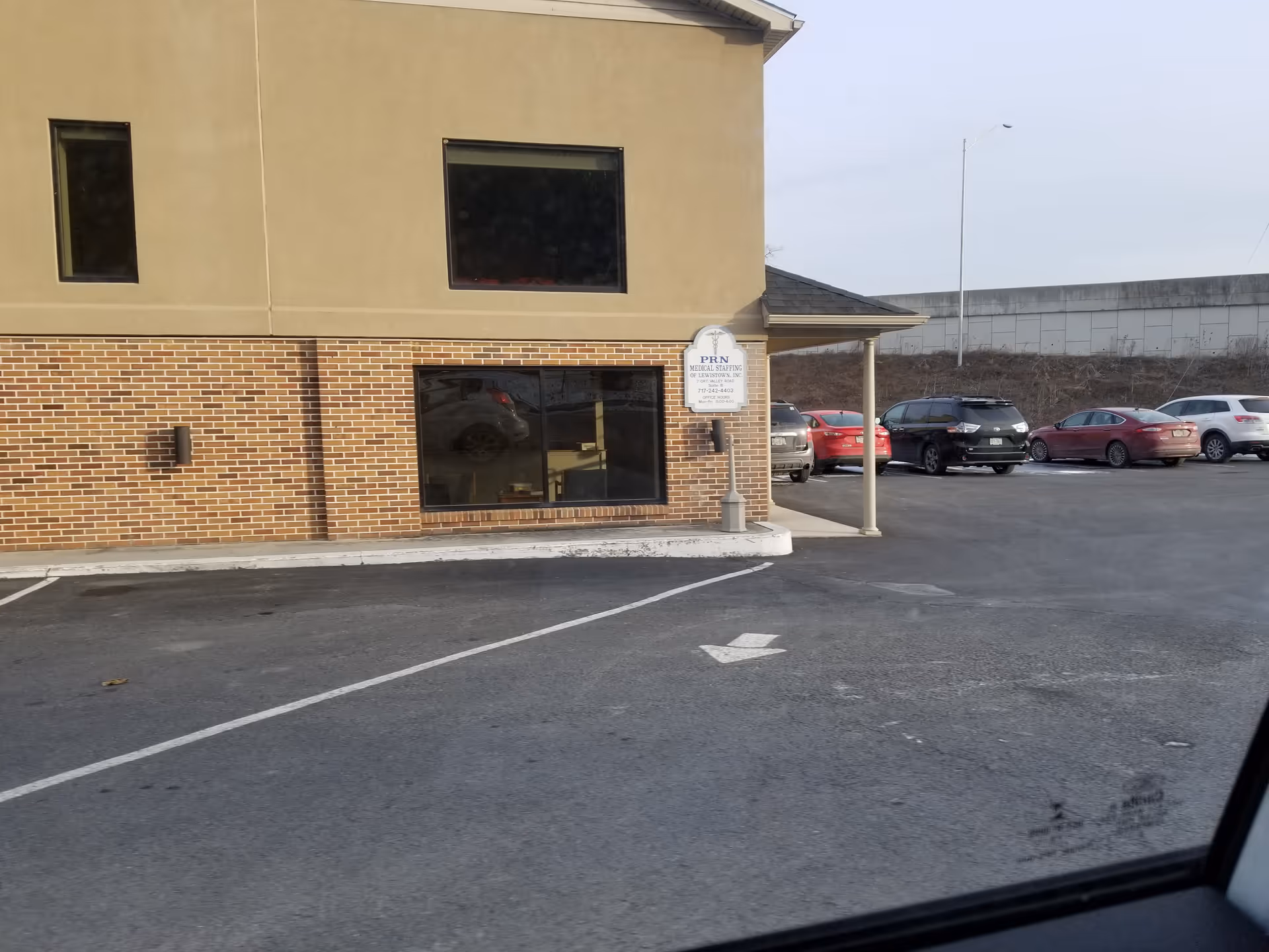 Exterior view of a tan-brick building with large windows, a sign, and a parking lot.