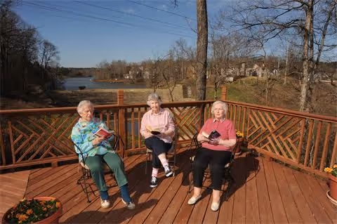 Three elderly women sitting on a wooden deck overlooking a lake, each holding a book.