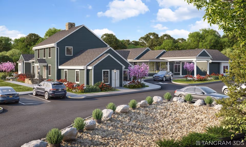 Exterior view of a senior living facility with multiple connected buildings painted in dark green with white trim. The scene includes a parking lot with several cars, landscaped areas with rocks, shrubs, and flowering trees, and a clear blue sky with some clouds.