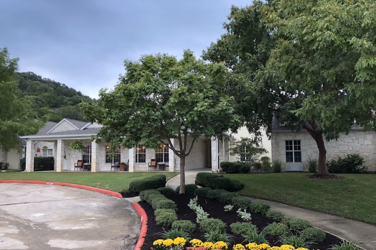 Exterior view of New Haven Kerrville facility showing a stone building with a covered porch, surrounded by green trees, shrubs, and a flower bed with yellow flowers. The sky is overcast.