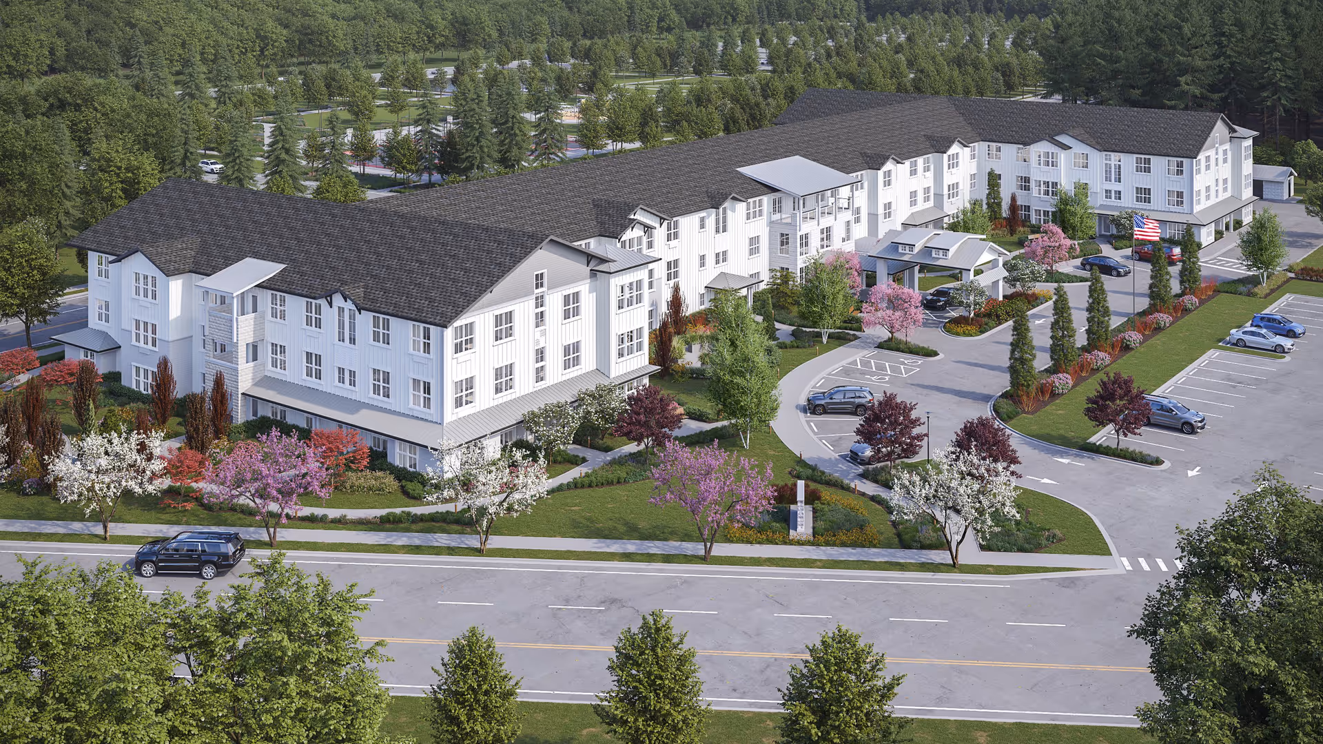 Aerial view of a large, modern senior living facility named The Esther at Riverbend, surrounded by landscaped gardens with colorful trees and shrubs. The building is three stories tall with white siding and dark roofing. There is a parking lot with several cars and an American flag near the entrance. The facility is set in a green area with many trees and a road in the foreground.