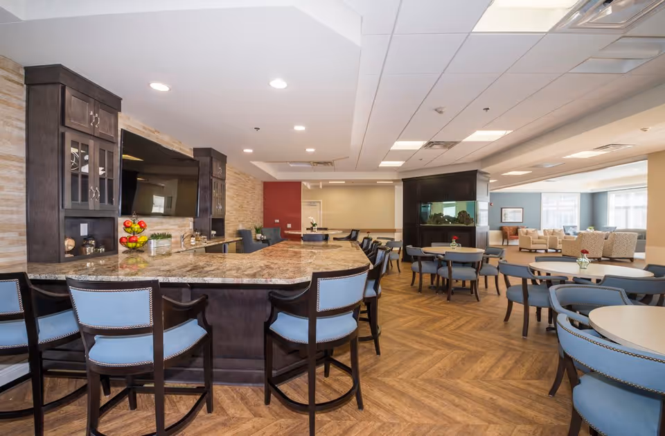 A spacious and well-lit common area in a senior living facility featuring a large granite countertop bar with blue cushioned chairs, round tables with blue chairs, a large flat-screen TV mounted on a wall with wooden cabinetry, and a seating area with beige armchairs near large windows. The room has wood-patterned flooring and recessed lighting in the ceiling.