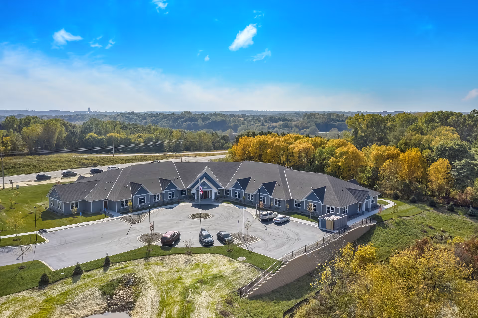 Aerial view of a single-story senior living building with a circular driveway, parked cars, and surrounding trees under a bright blue sky.