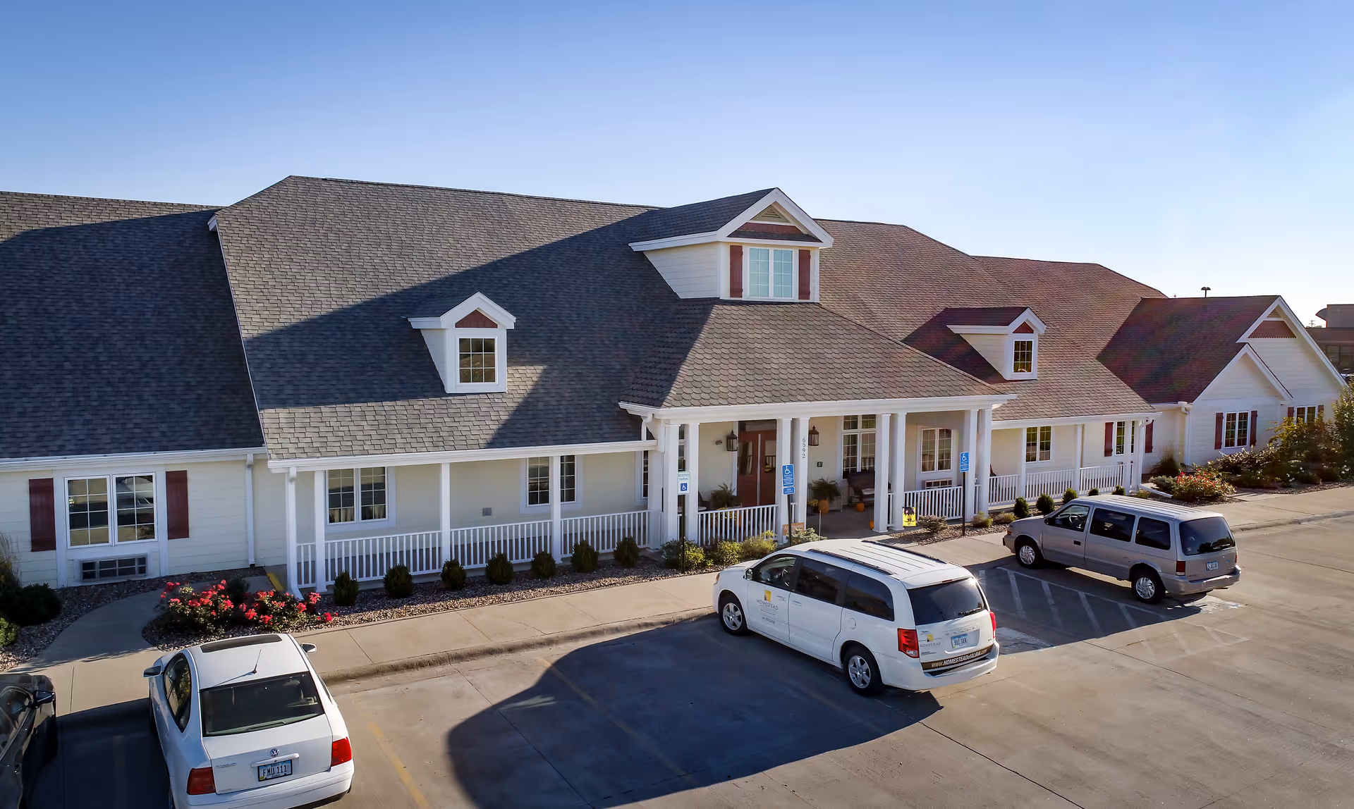 Exterior view of a single-story assisted living facility building with a large covered porch, multiple windows, and a sloped roof with dormer windows. There are three vehicles parked in front of the building in a paved parking lot, with some landscaping including bushes and flowers near the building.