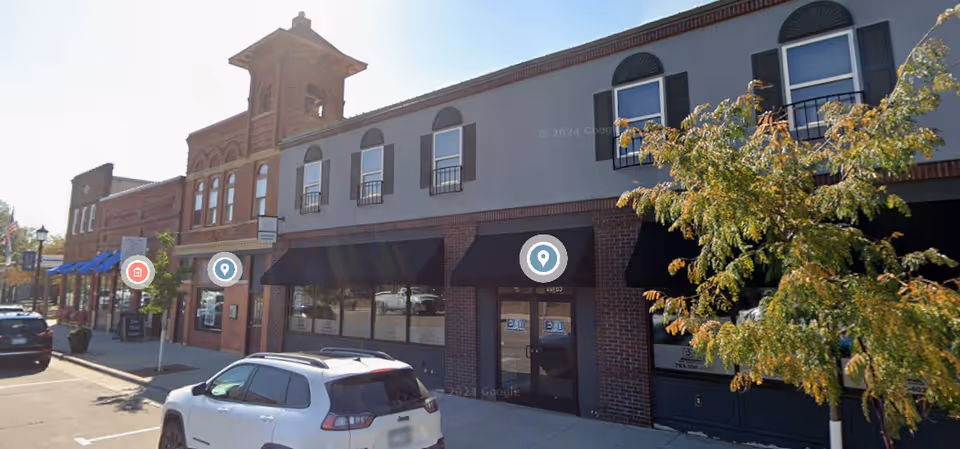 Exterior street view of brick storefronts with black awnings and parked cars.