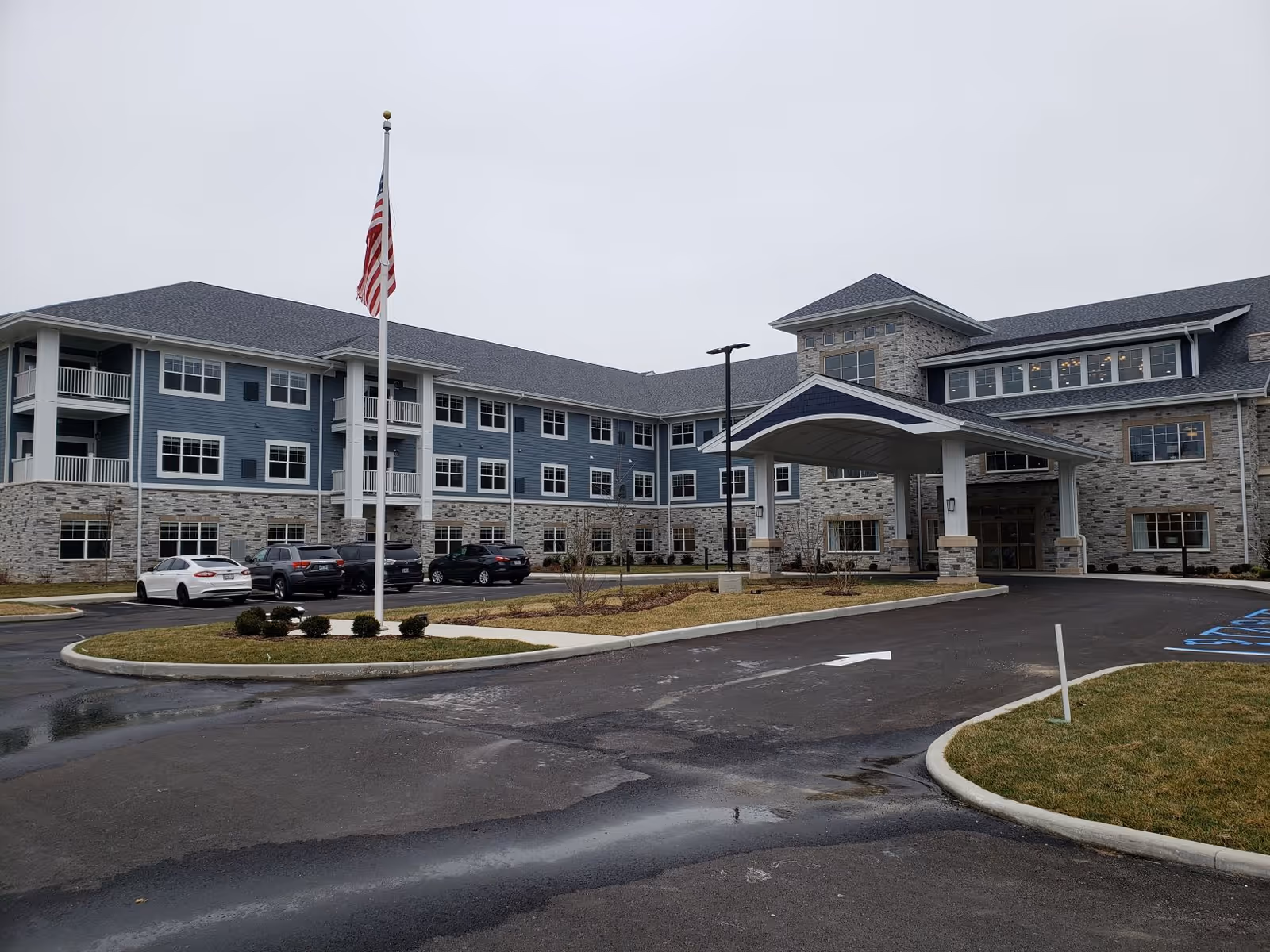 Exterior view of a large senior living facility building with a covered entrance, multiple windows, and a flagpole with an American flag in front. Several cars are parked in the parking lot, and the sky is overcast.