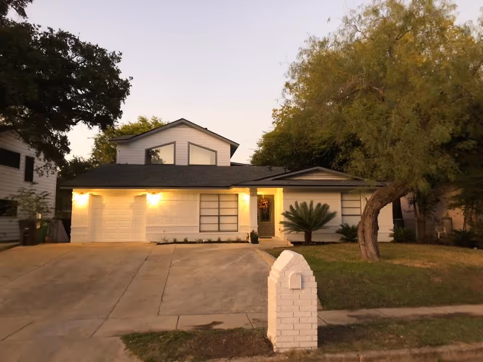 Two-story house front at dusk with lit exterior lights, a wide driveway, a white brick mailbox, and surrounding trees.