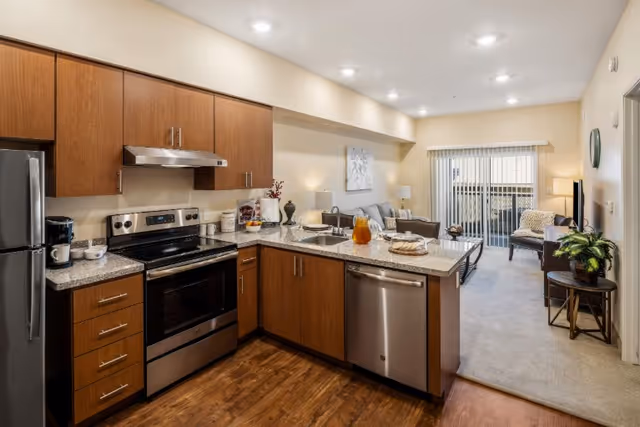 Open-concept kitchen with wooden cabinets, stainless steel appliances, granite countertops, and living area beyond.