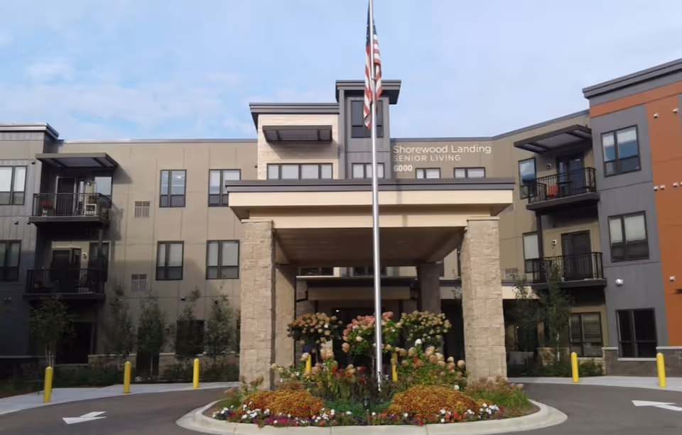 Front exterior view of Shorewood Landing senior living facility with a covered entrance, an American flag on a flagpole, and landscaped flower beds in front of the building.